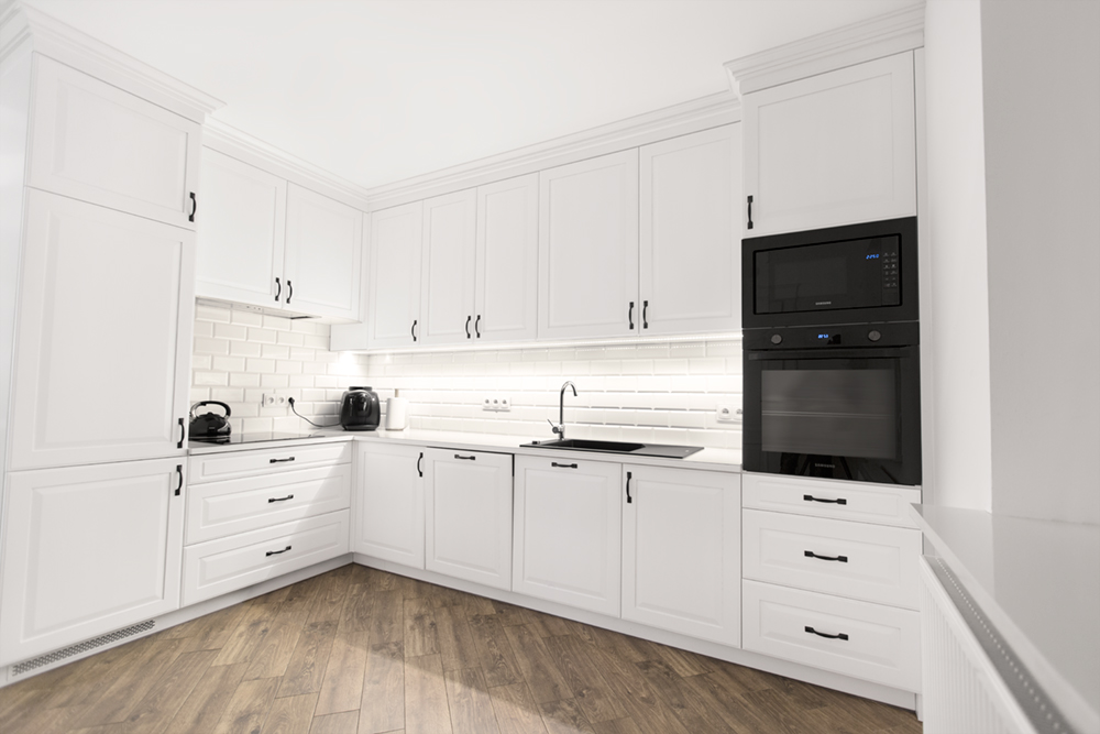 jasna biała kuchnia w kształcie litery L z zabudową do sufitu, czarnym AGD i drewnianą podłogą – fotografia wnętrz kuchni na wymiar Kulenty